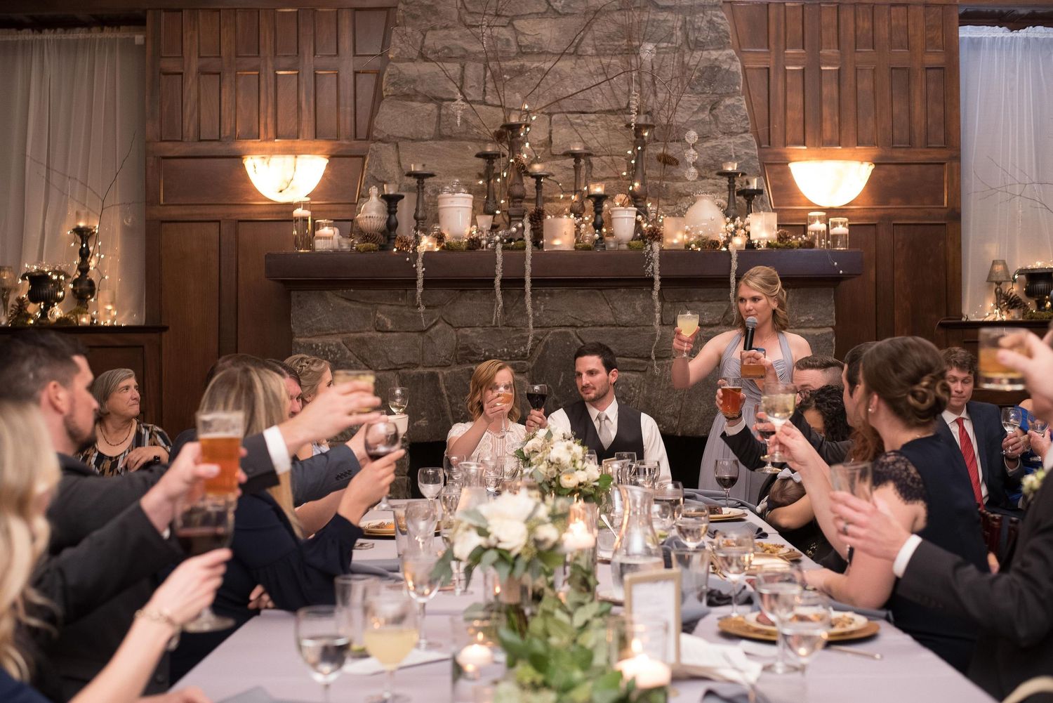 Wedding guests raise glasses in a toast at an elegant reception with a stone fireplace and warm chandelier lighting.