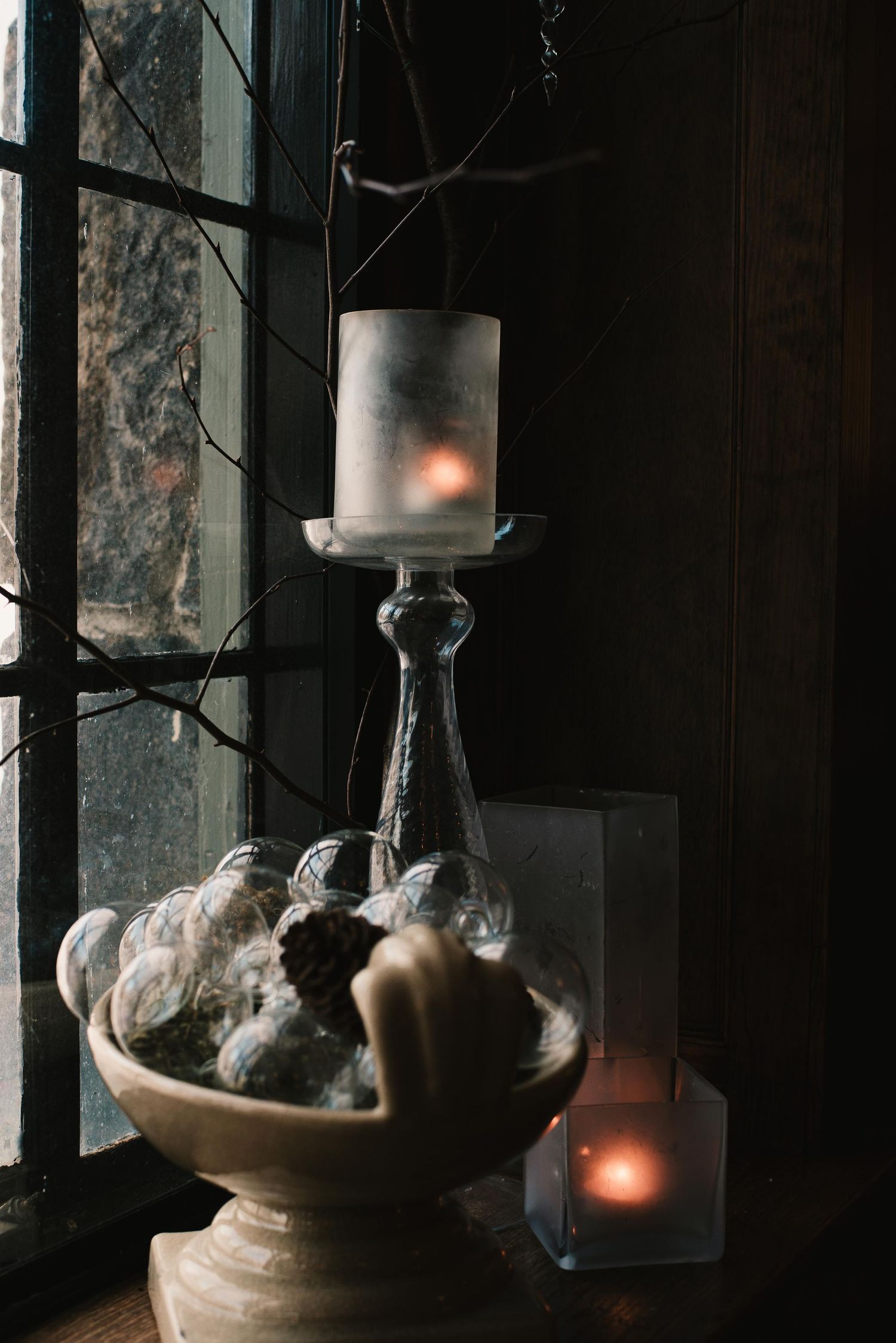 A lit pillar candle on a glass candlestick beside a ceramic bowl with decorative objects near a dark window.