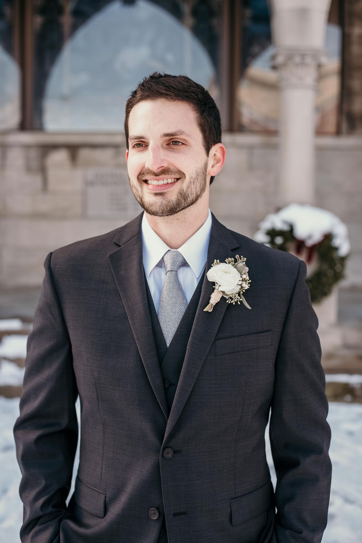 Smiling groom in dark suit with white boutonniere and gray tie posing outdoors at winter wedding venue.