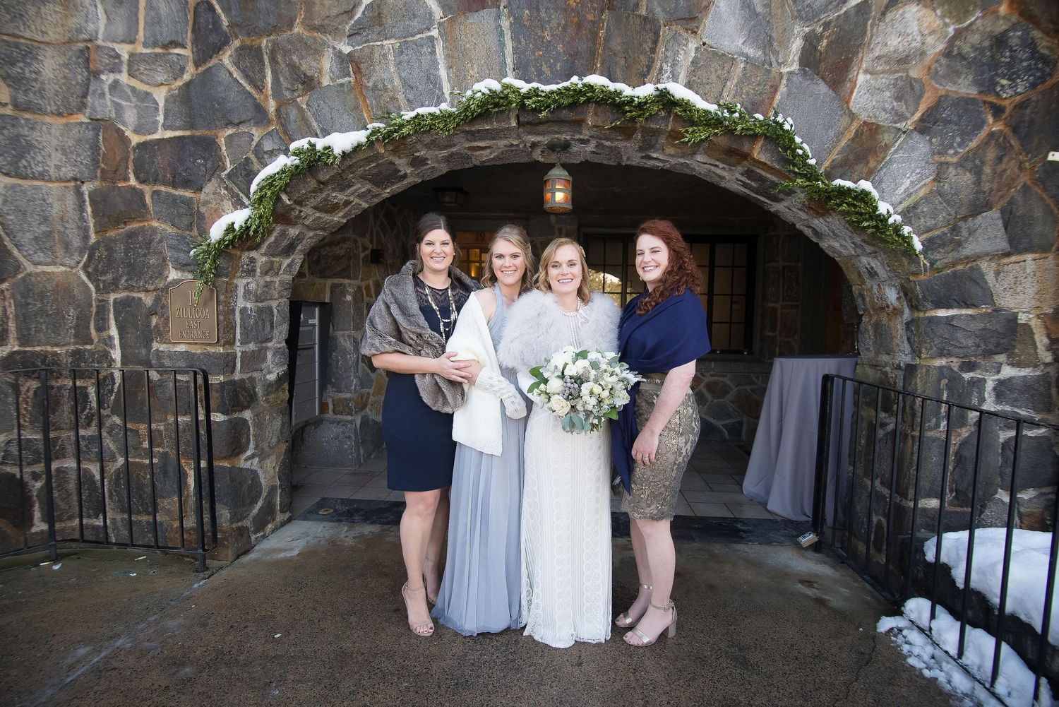 Bride with bouquet and bridesmaids in navy and gray dresses pose under stone archway with greenery at winter wedding.