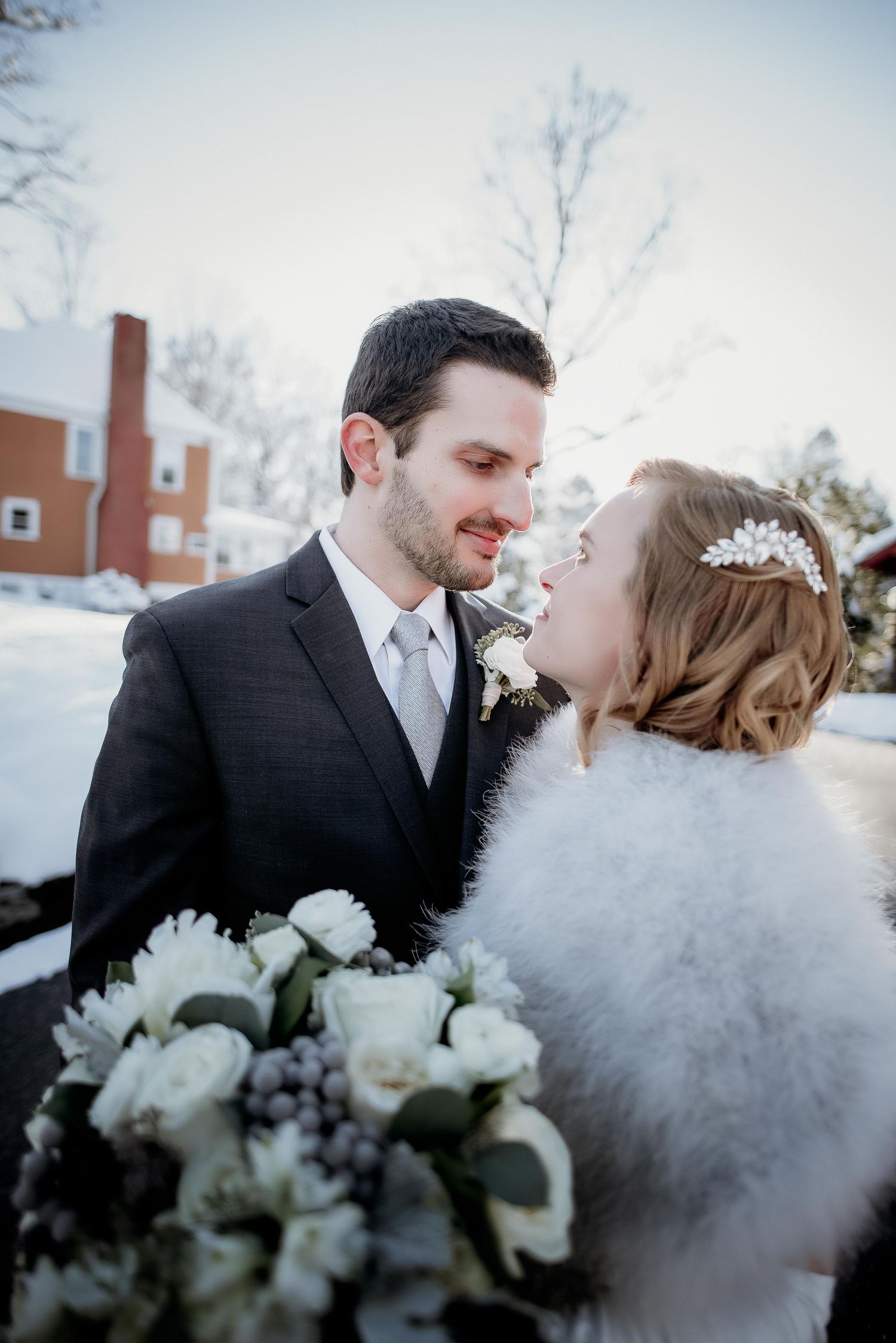 Bride and groom share a romantic winter moment, she holds white roses bouquet, wearing fur wrap and floral headpiece.