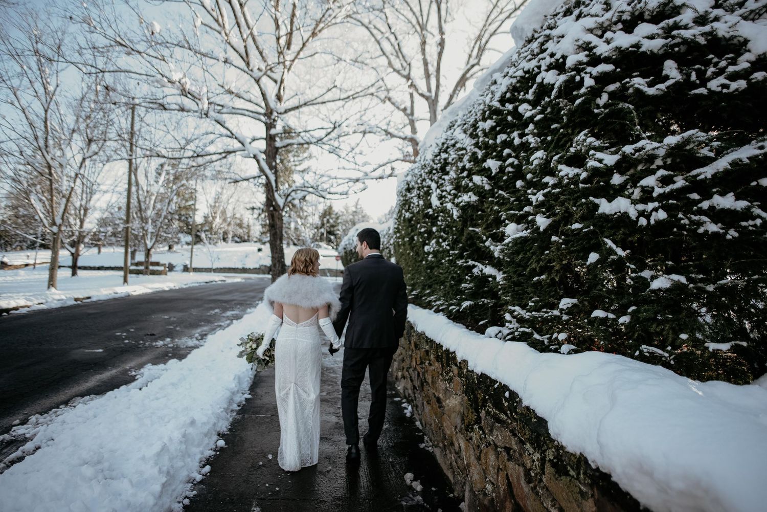 Bride and groom walk hand in hand along a snowy path beside a stone wall and snow-covered trees in winter.