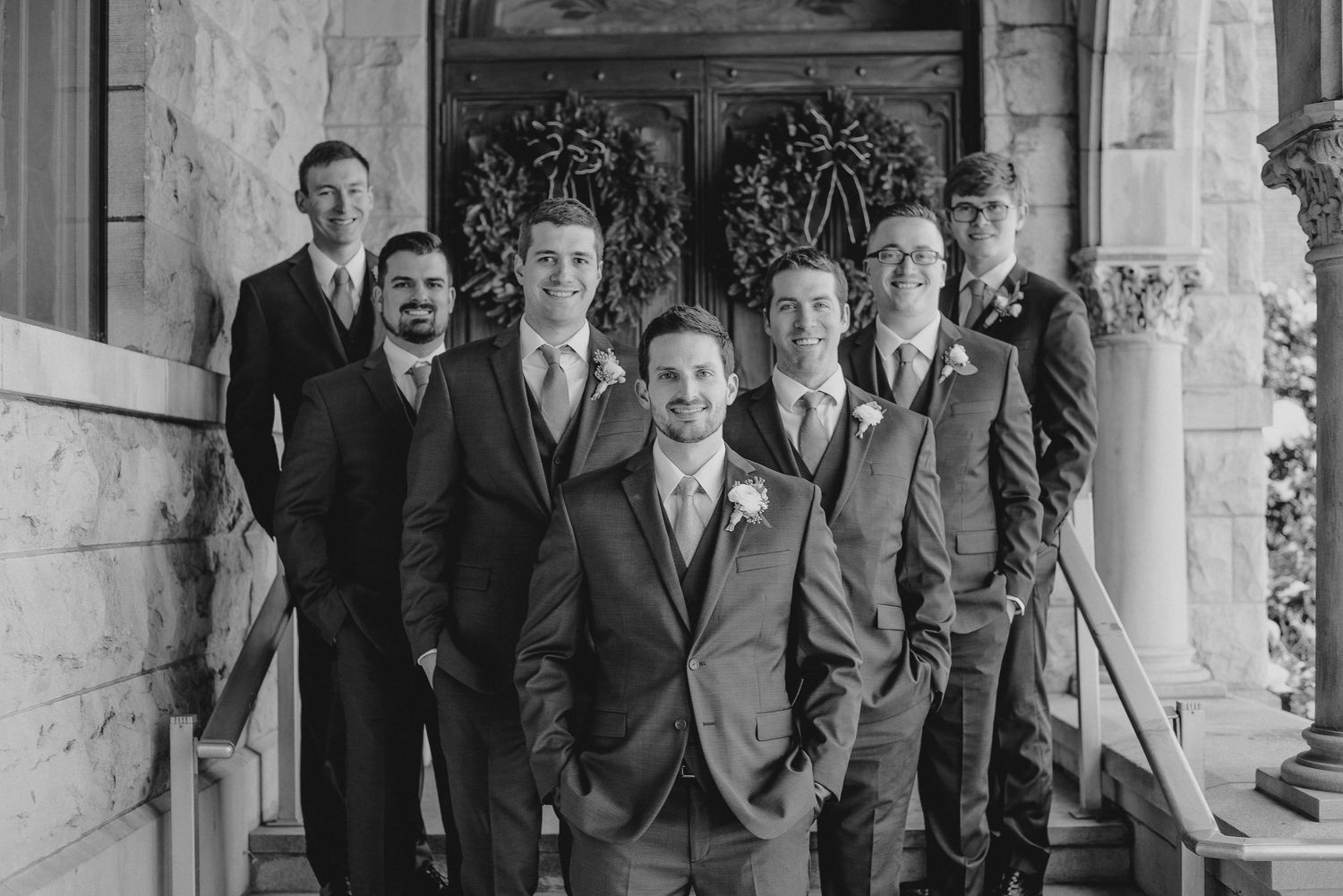 Black and white photo of a groom and groomsmen in suits posing on church steps for a wedding portrait.