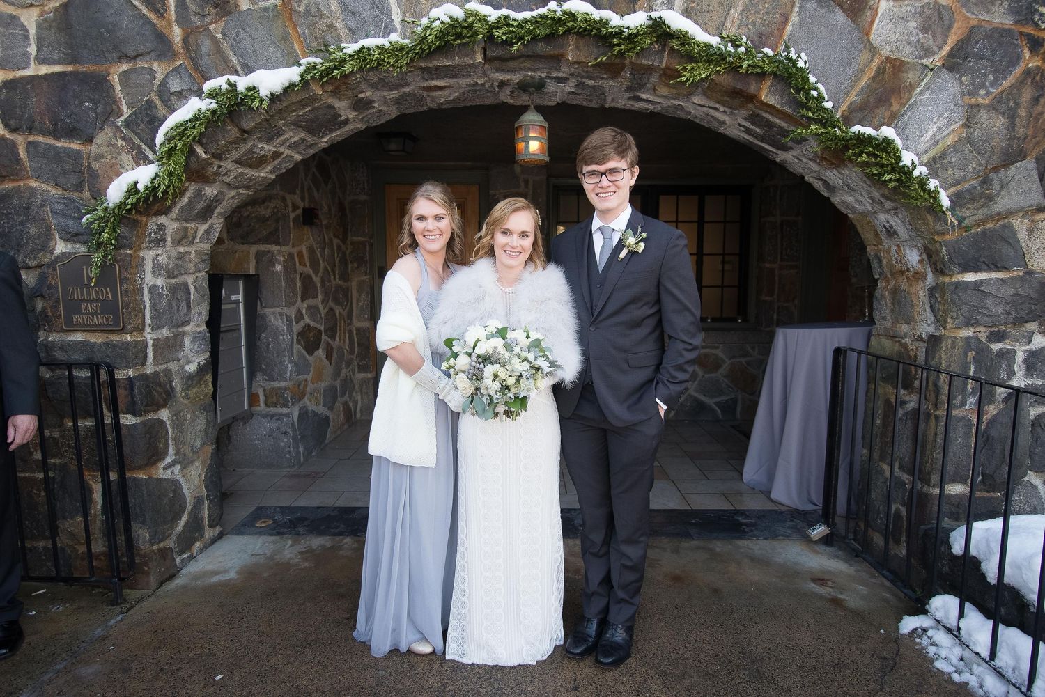 Bride in white gown with bridesmaid in blue and groomsman in black suit posing under stone arch in winter wedding.