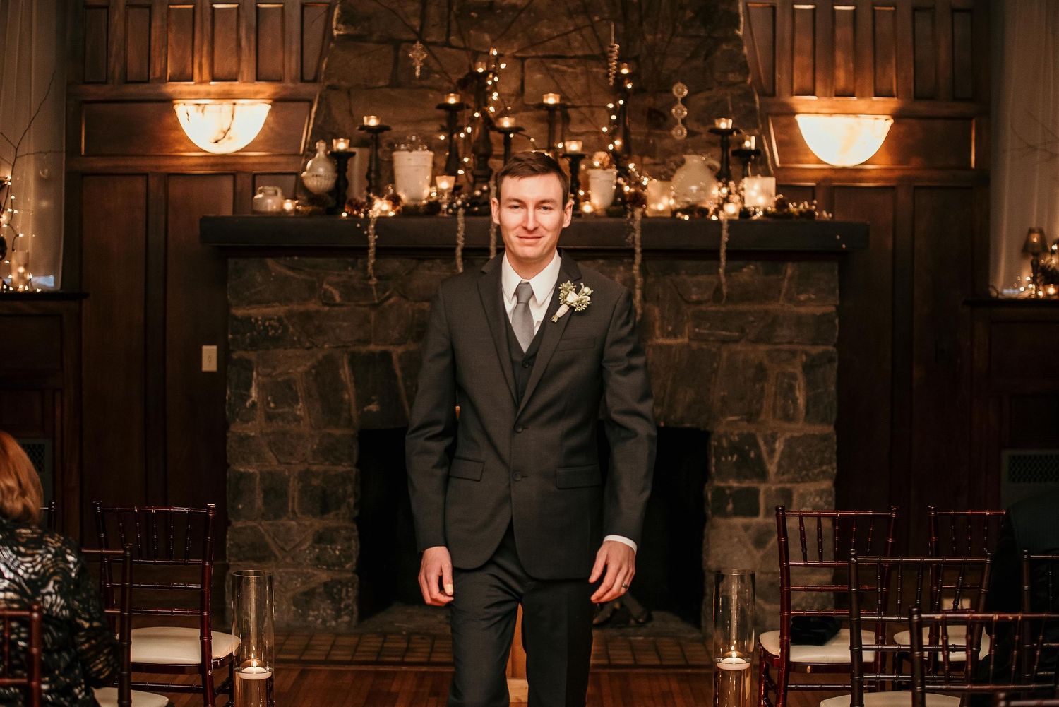 Groom in dark suit with boutonniere stands before stone fireplace in elegant wedding venue.