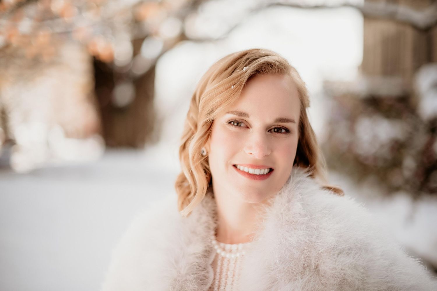 Smiling blonde woman in a white fluffy wrap poses outdoors in a snowy winter setting with bokeh lights.