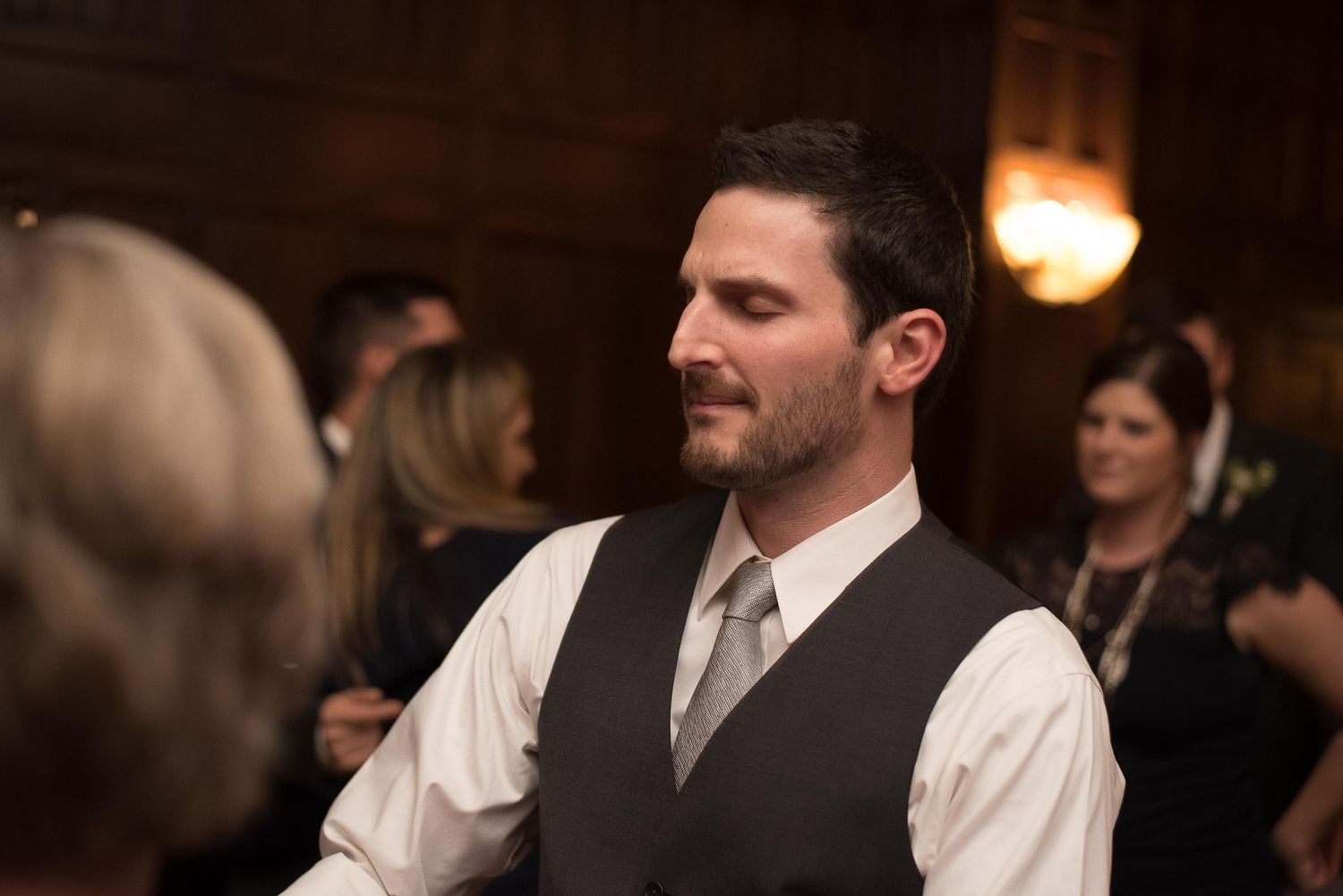 A well-dressed man in a vest and tie smiles at a formal indoor event or wedding reception.