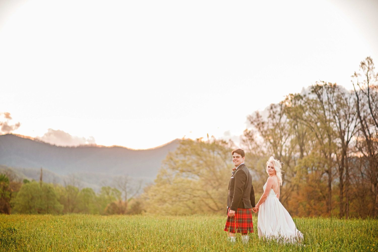 Bride and groom in Scottish kilt holding hands in green meadow with mountain backdrop at golden hour.