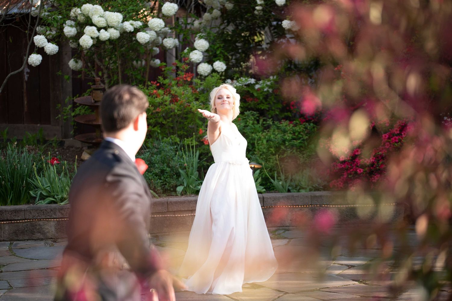 Bride in white gown reaches out to groom in garden with blooming flowers during outdoor wedding ceremony.