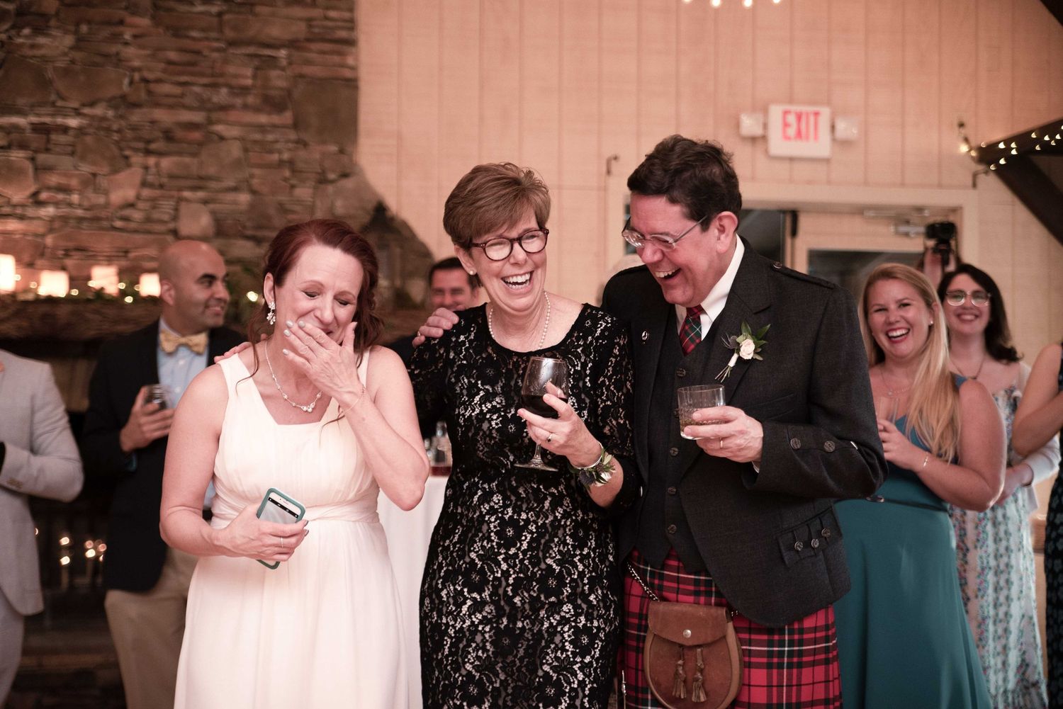 Joyful wedding reception with bride in white, woman in black lace, and man in red tartan kilt laughing together.