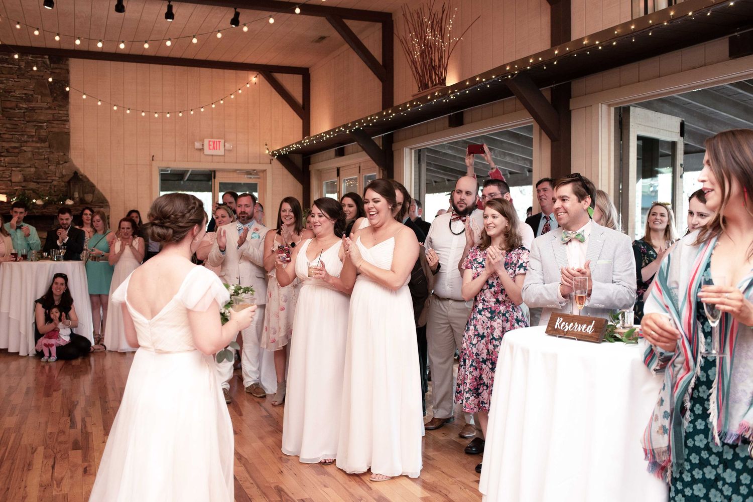 Bride tosses bouquet to bridesmaids in white dresses at elegant barn wedding reception with string lights.
