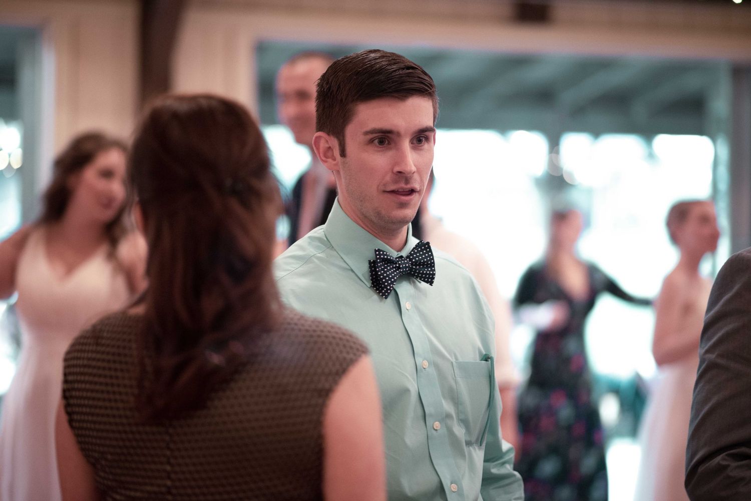 Young man in bow tie and mint shirt dancing at a wedding reception with guests in background.