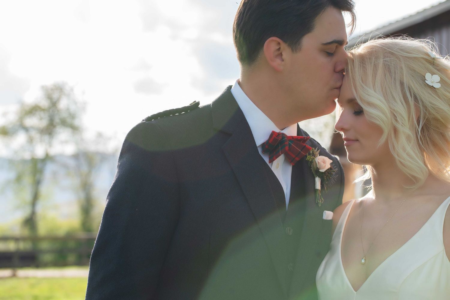 A groom in a navy suit with red bow tie kisses his blonde bride on the forehead outdoors on their wedding day.