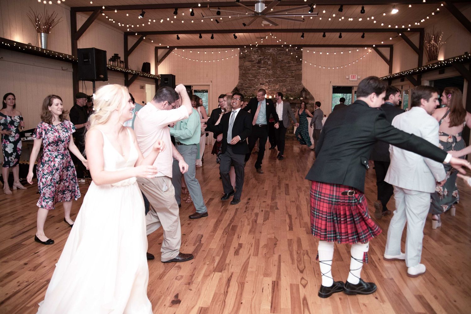 Wedding guests dancing joyfully on a wooden dance floor, including a man in a Scottish kilt, at a rustic barn reception.