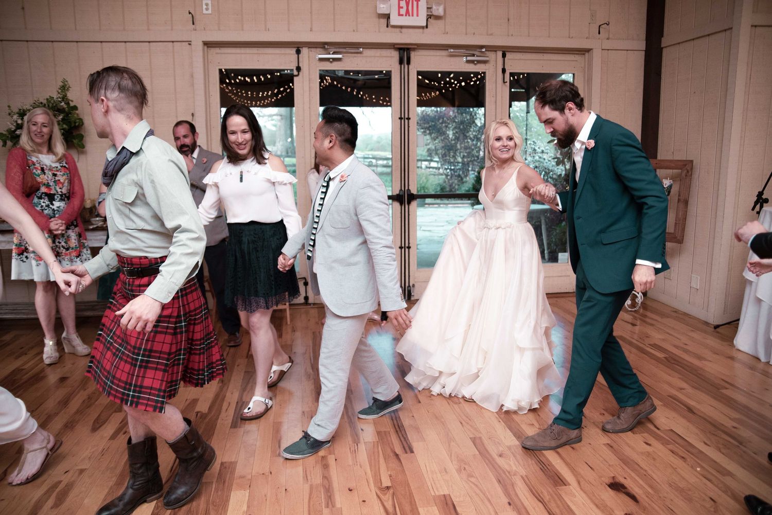 Wedding guests dancing joyfully on wooden dance floor, bride in white gown with groom in green suit at reception.