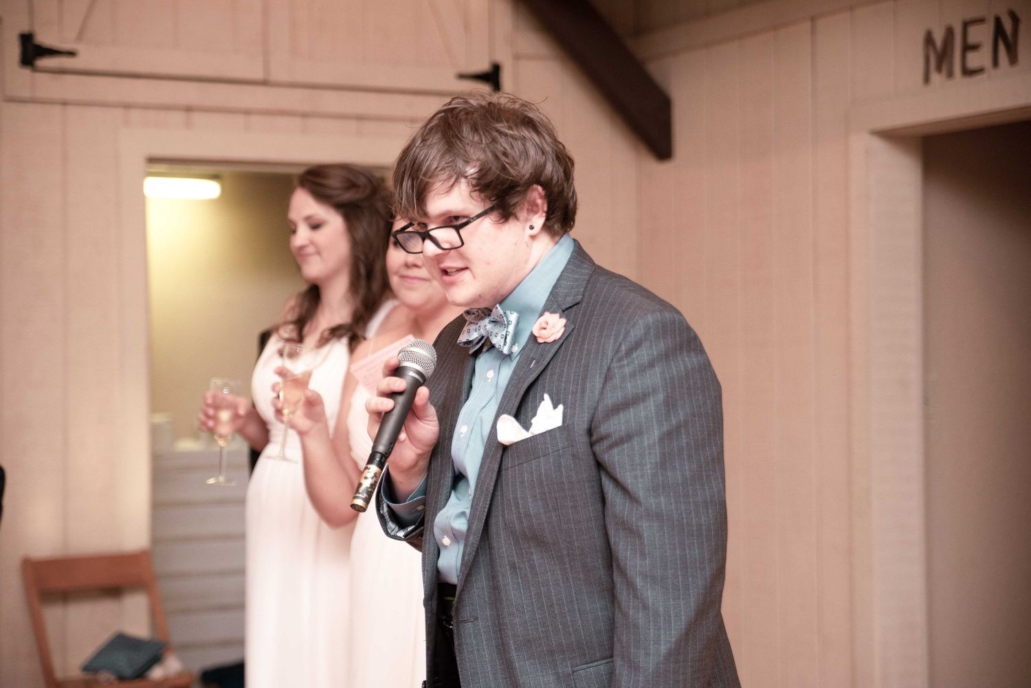 Person in gray suit speaking into microphone at wedding reception with woman in pink dress in background.