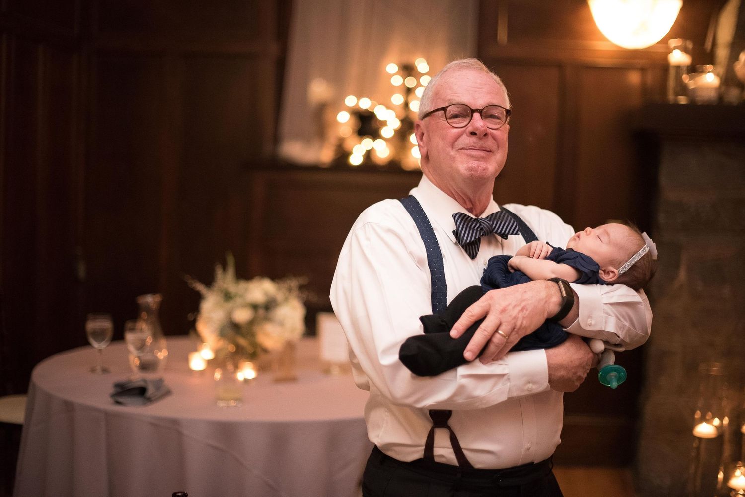 Elderly man in suspenders and bow tie holding a newborn baby at an elegant wedding reception.
