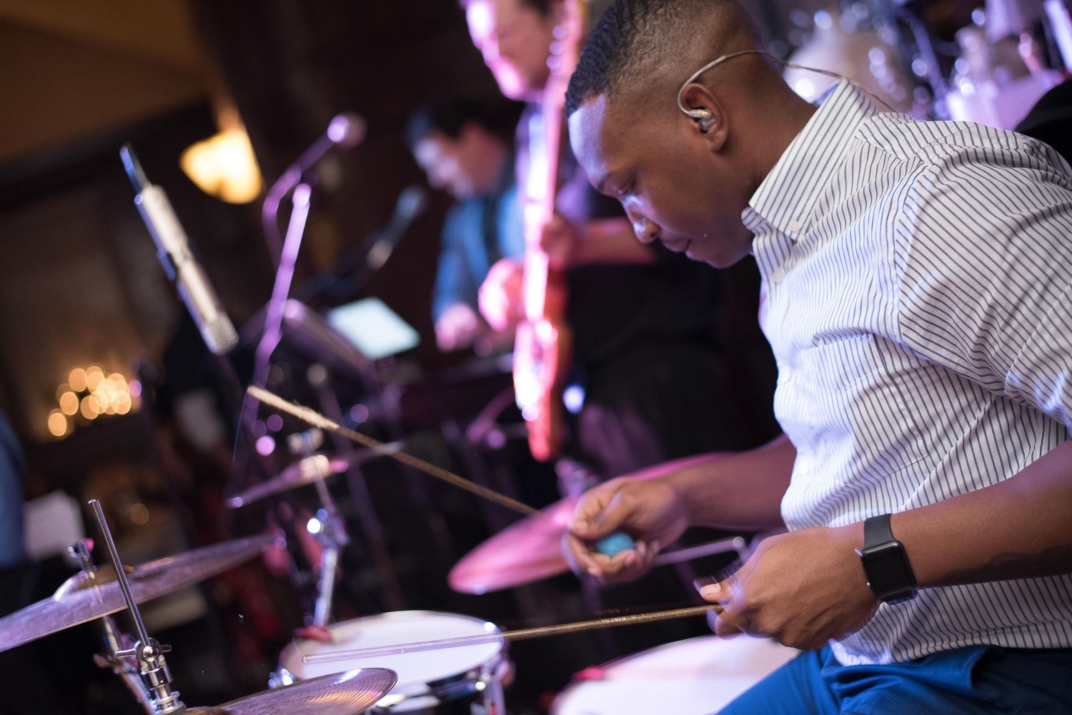 A musician plays drums with drumsticks at a live band performance in a dimly lit venue.