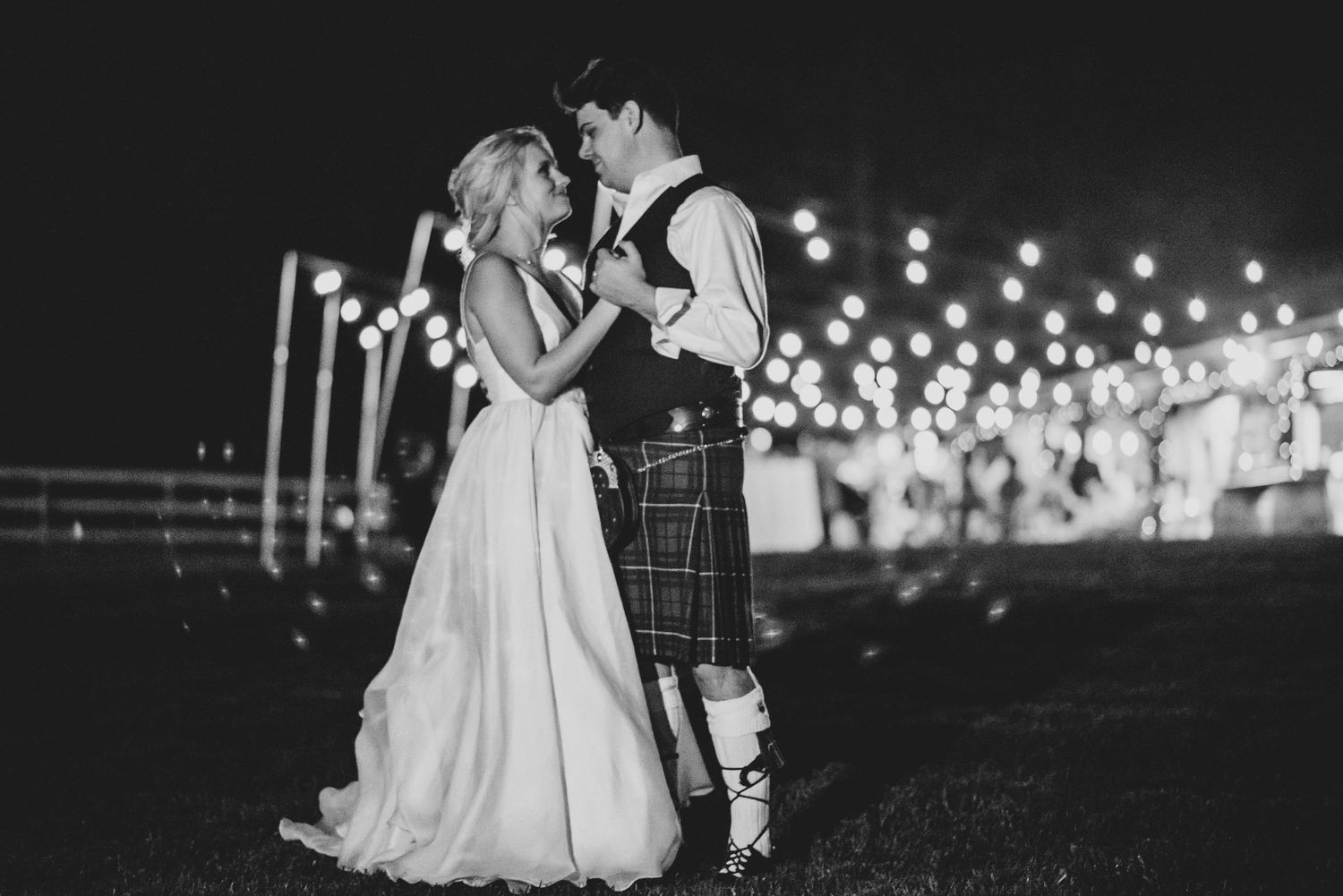 Black and white photo of a bride and groom sharing a romantic first dance outdoors under glowing lights at night.