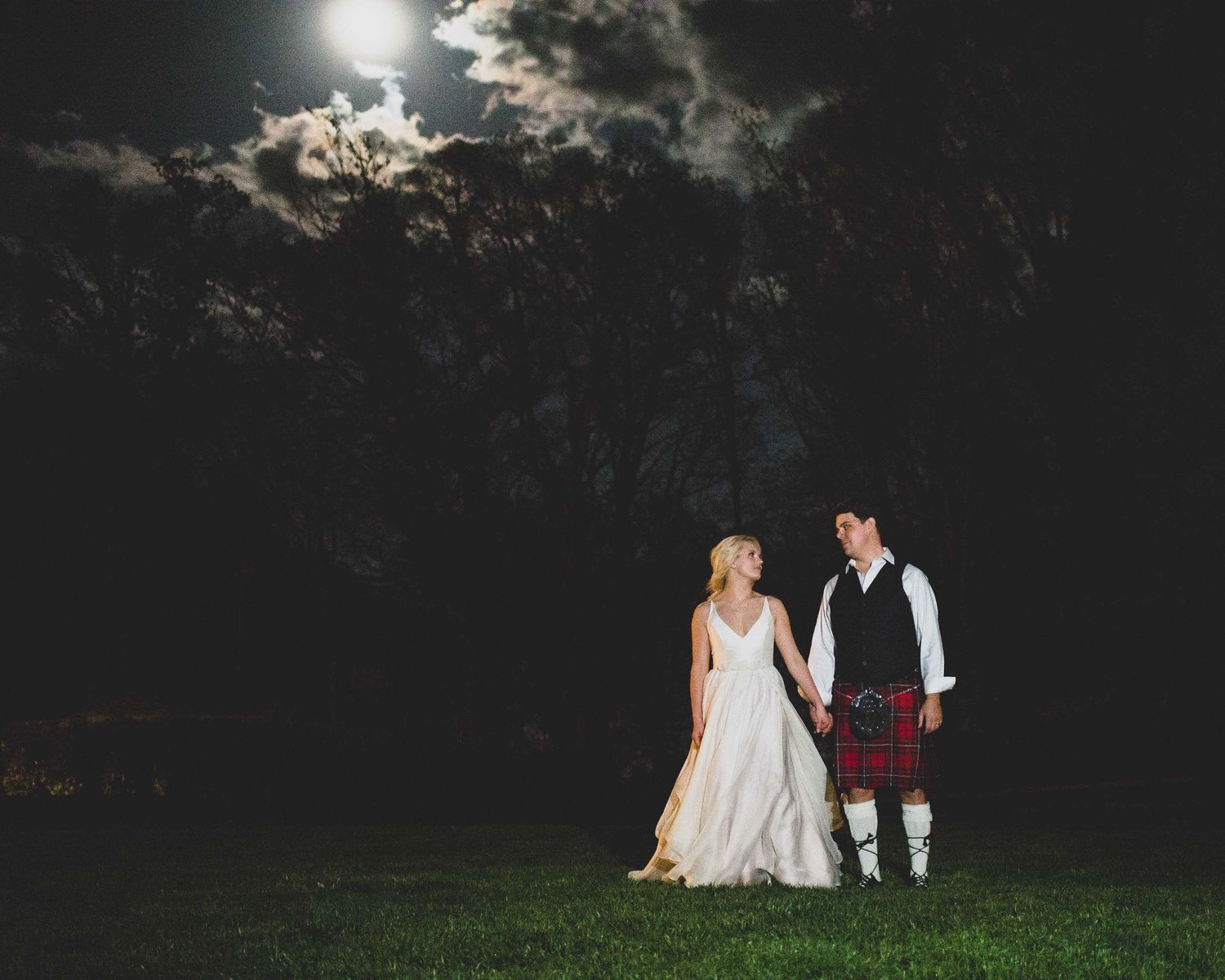 Bride and groom in Scottish kilt stand romantically under moonlit night sky at outdoor wedding.