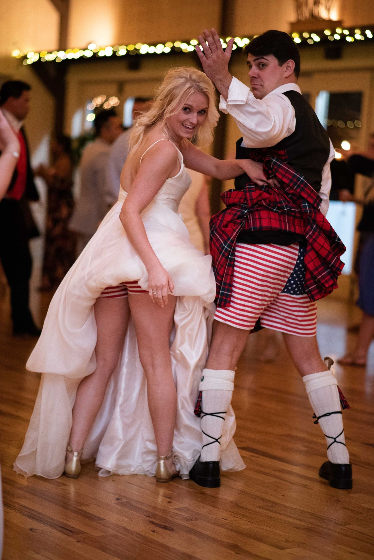 Bride in white gown and man in tartan kilt dancing energetically at a wedding reception on a wooden dance floor.