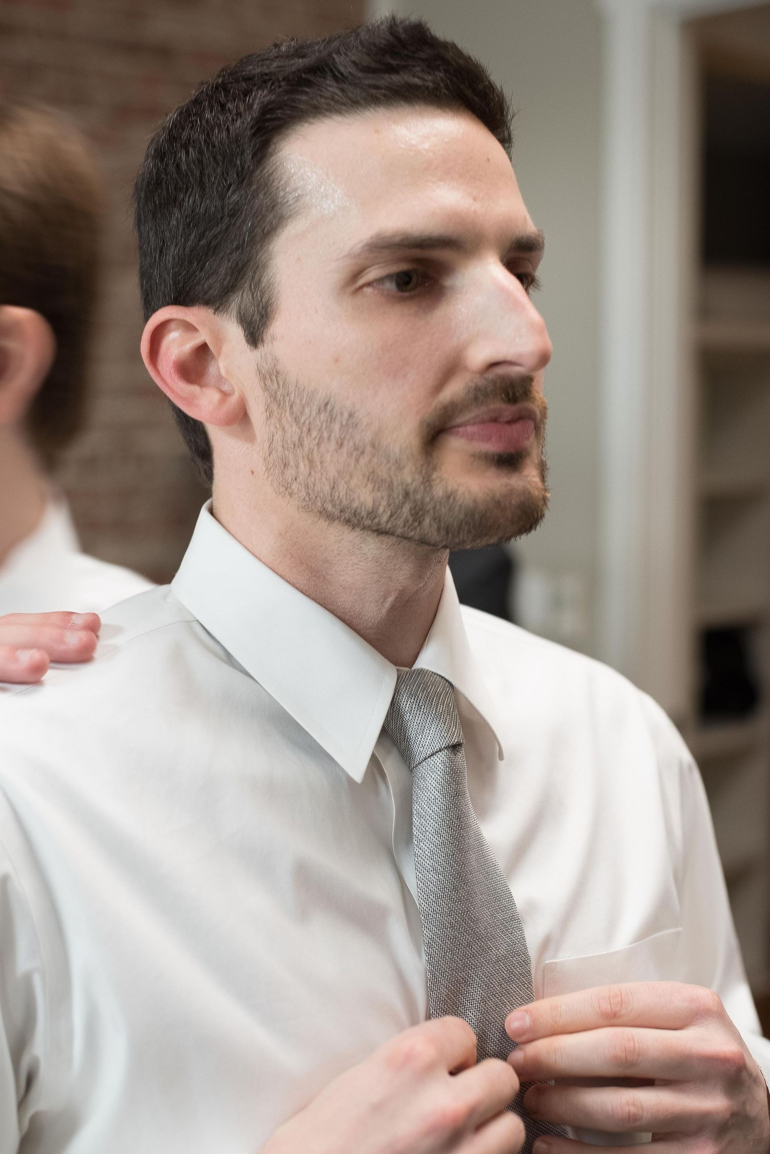 Man in white dress shirt adjusting gray tie, with another person's hand visible on his shoulder.
