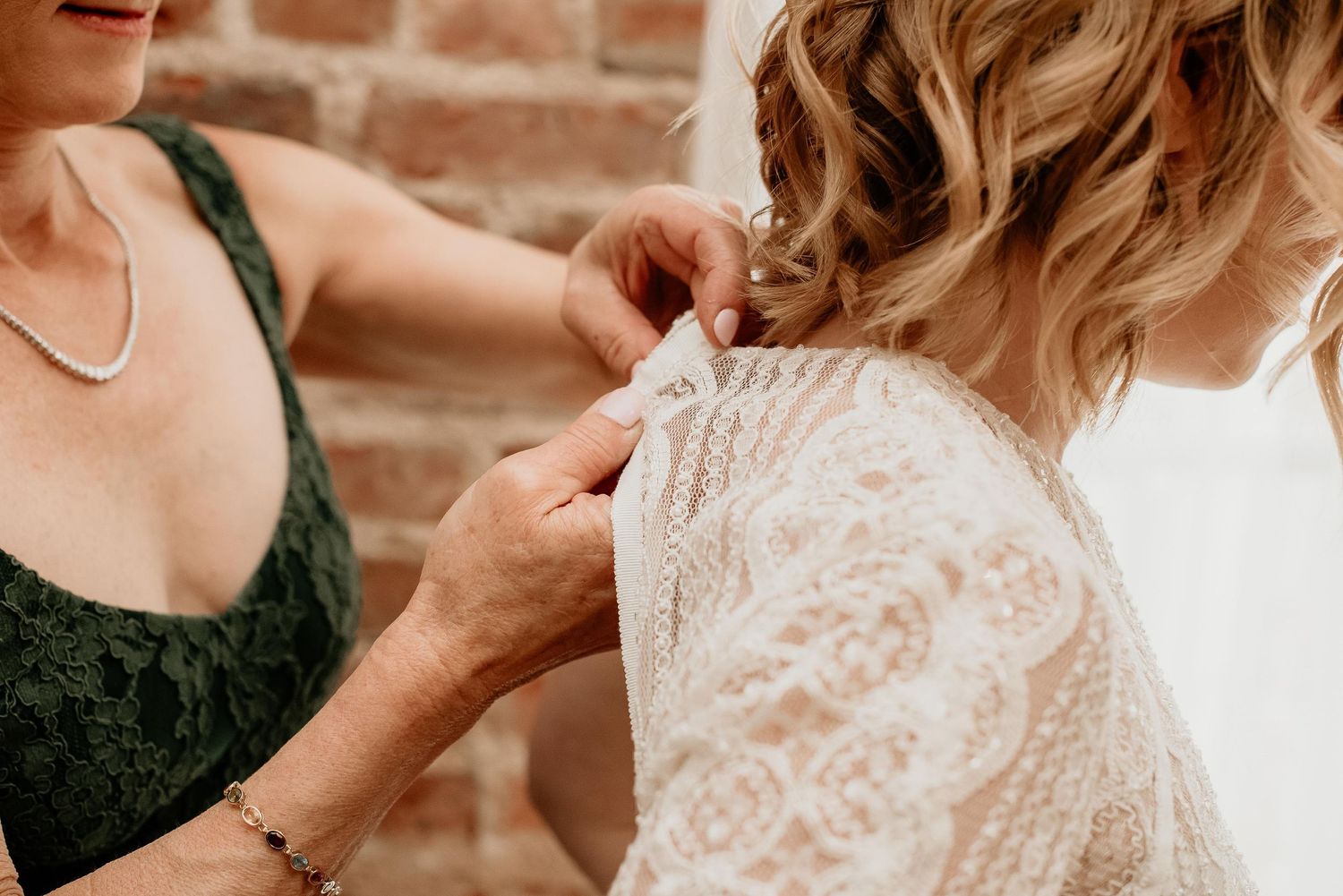 Woman in green lace dress buttoning the back of a bride's white lace wedding gown near a brick wall.