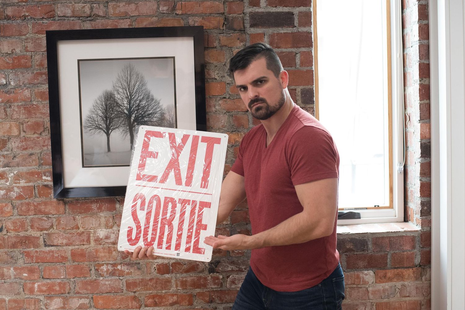 Man holding bilingual EXIT SORTIE sign against rustic brick wall with framed tree artwork in background.
