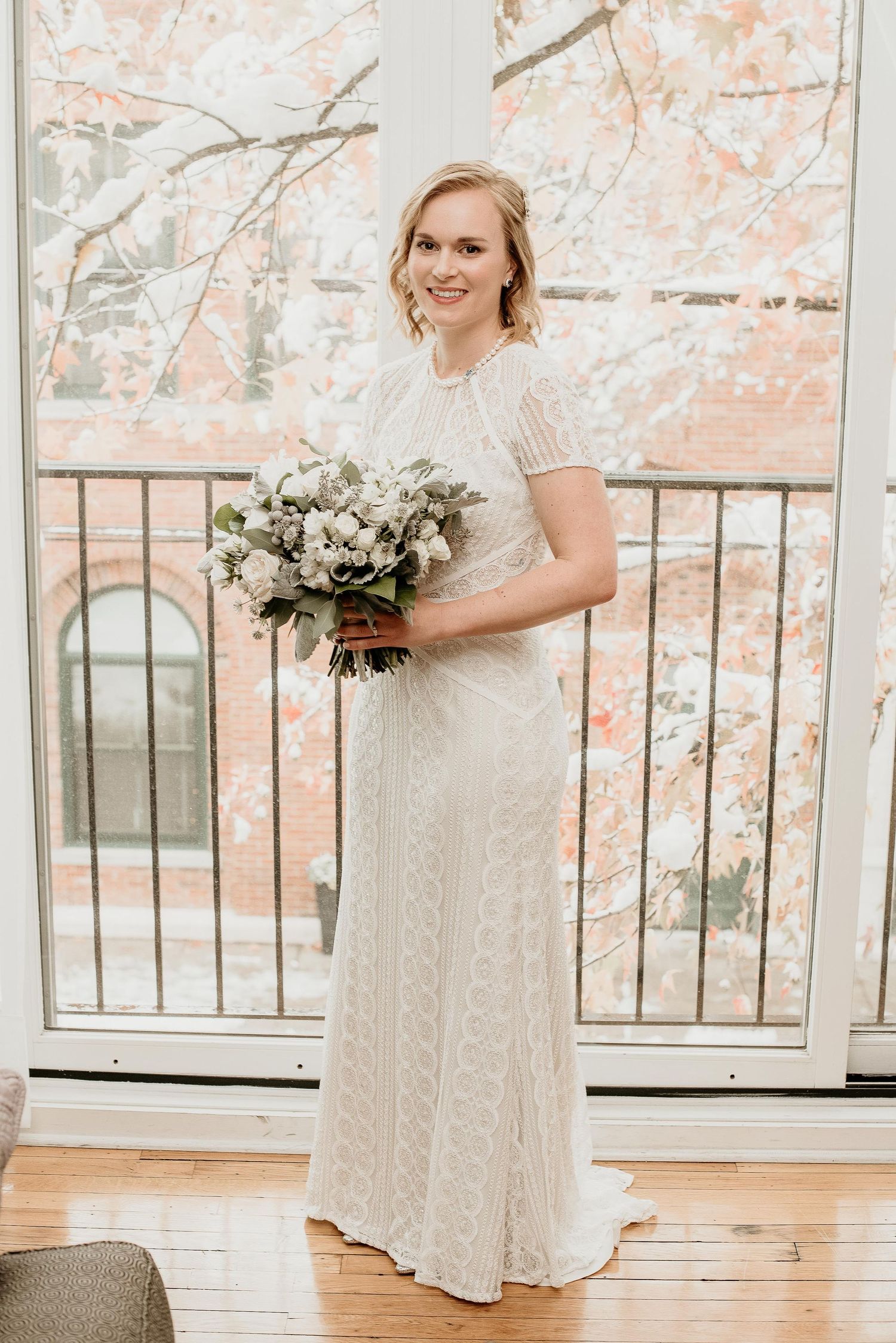 Bride in white lace gown holding bouquet stands before snowy window backdrop on her wedding day.