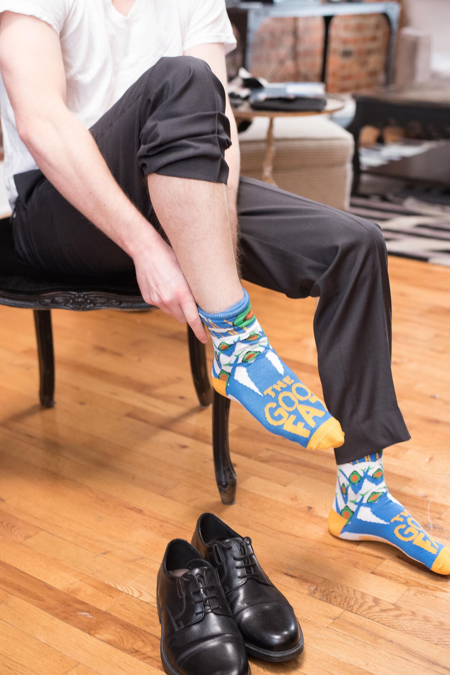Man putting on blue and yellow novelty socks with black dress shoes on hardwood floor nearby.