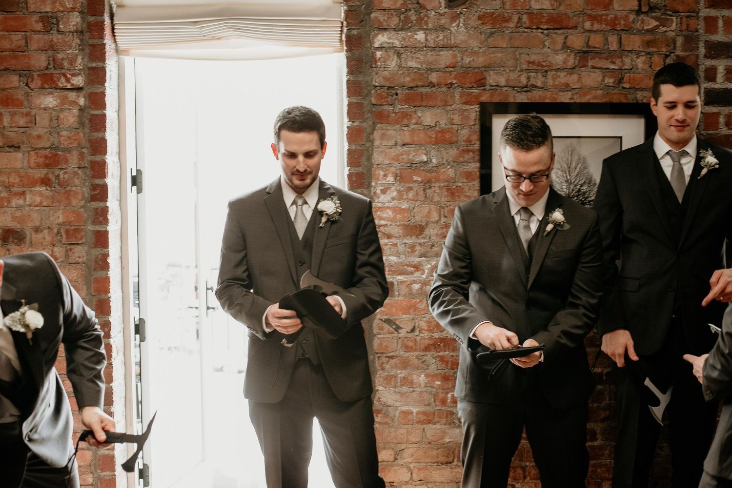 Groomsmen in gray suits with boutonnieres checking phones before a wedding ceremony in a brick-walled venue.