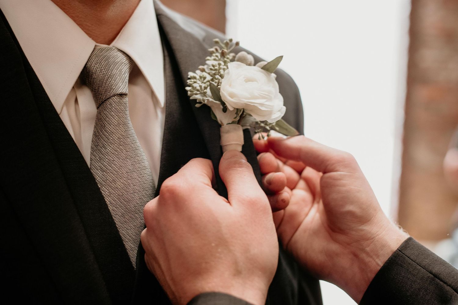 Hands pinning a white rose boutonniere onto a groom's dark suit lapel with a gray tie at a wedding.