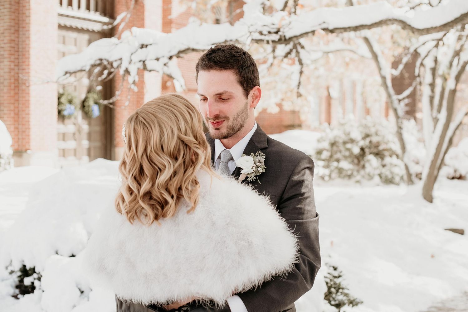 Bride in white fur wrap and groom in gray suit share an intimate moment outdoors in a snowy winter wedding setting.