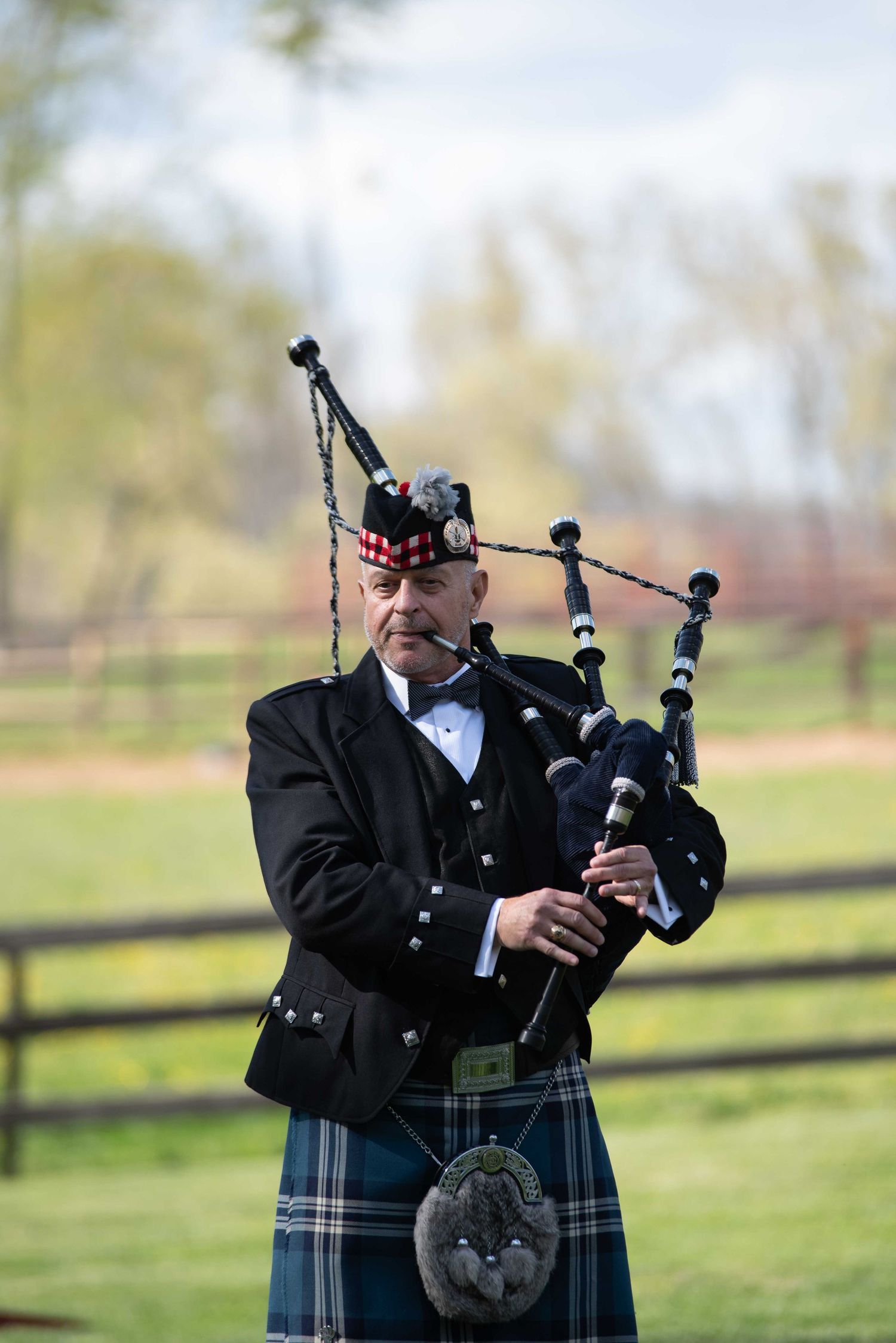 A Scottish bagpiper in traditional tartan kilt and black jacket performs outdoors on a green field.