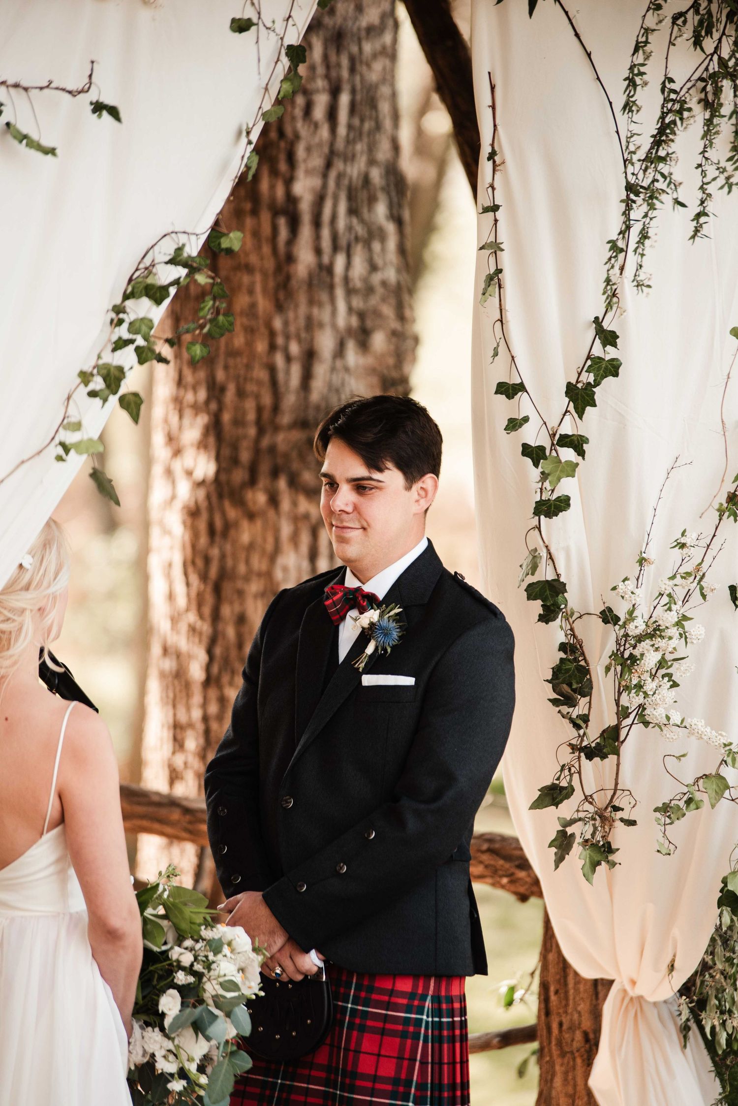 Groom in black tuxedo and red tartan kilt smiling at bride during outdoor woodland wedding ceremony.