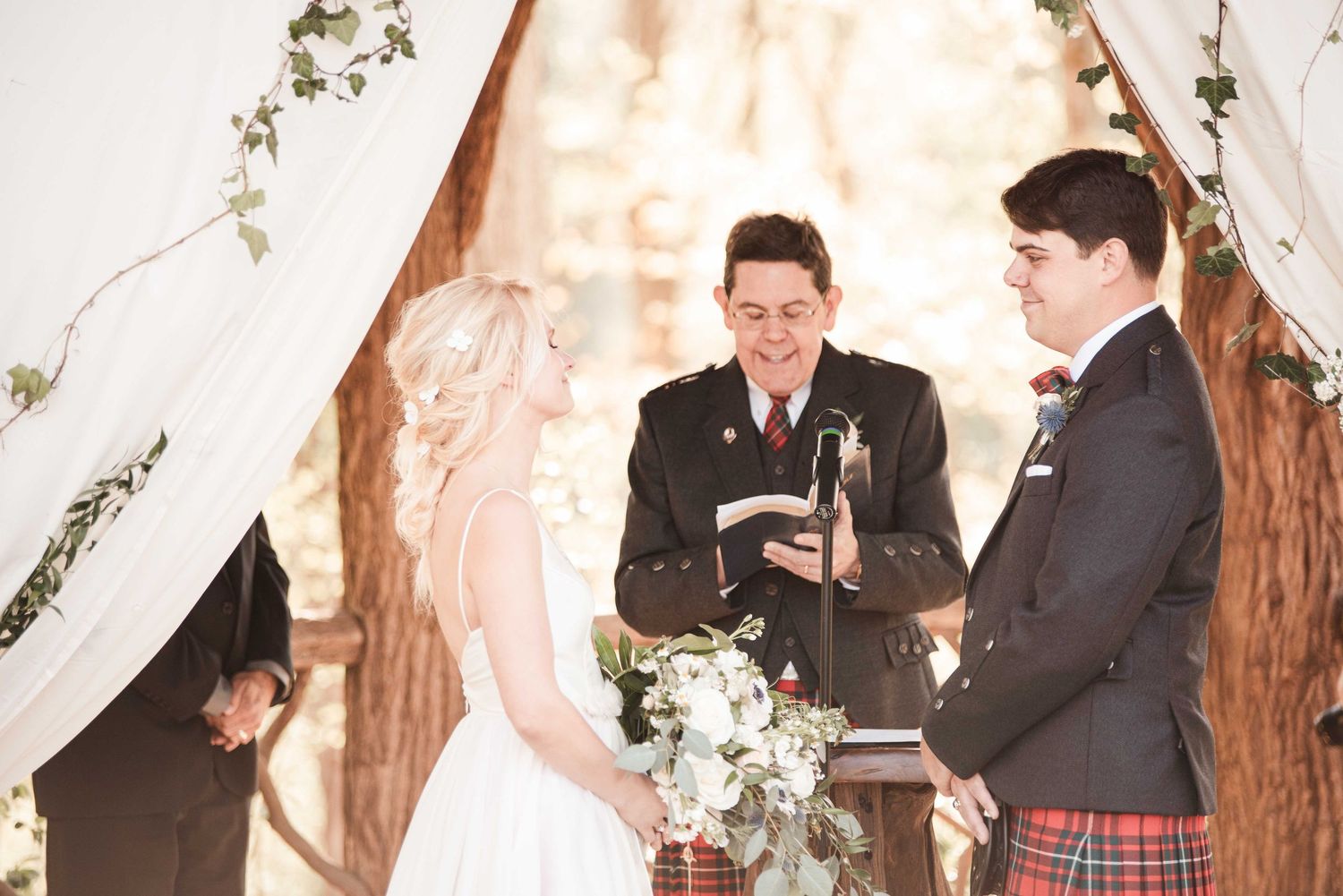 Outdoor wedding ceremony with bride, groom in tartan kilt, and officiant under floral arch among trees.