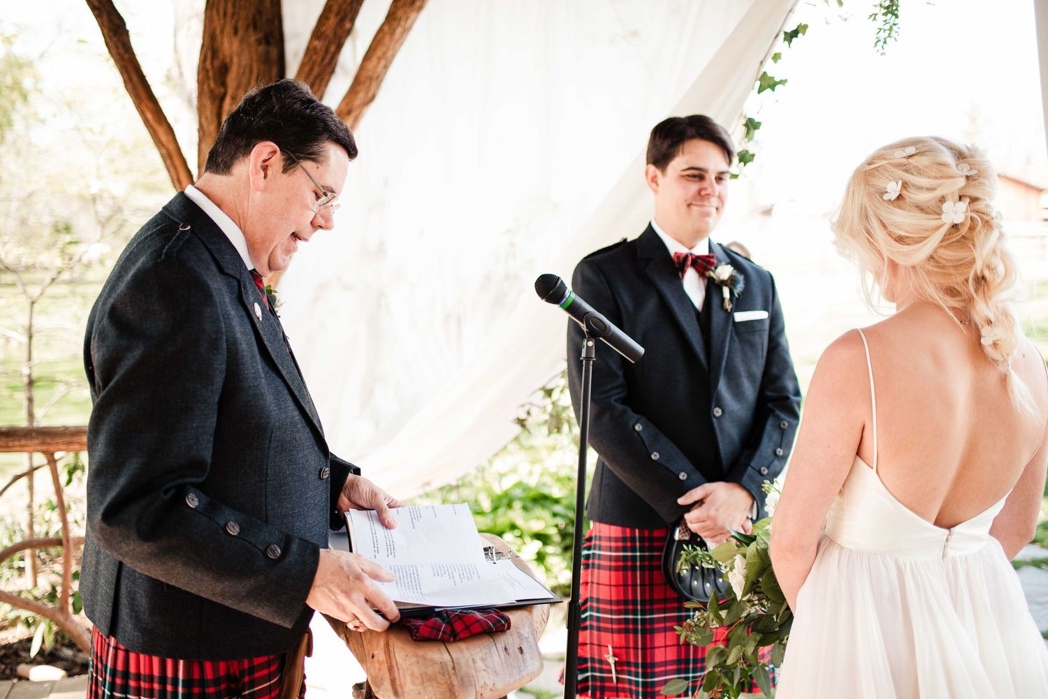 Wedding ceremony with groom in tartan kilt and officiant reading vows outdoors, bride in white dress facing them.