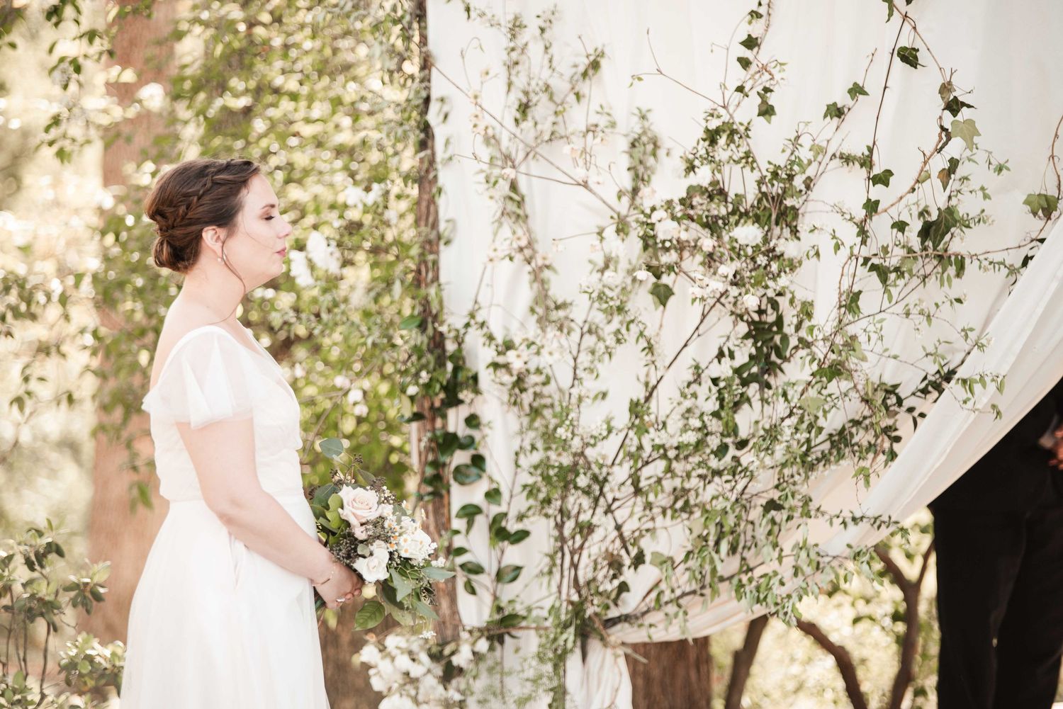 Bride in white gown holding bouquet stands beside flowering branches and draped fabric backdrop outdoors.