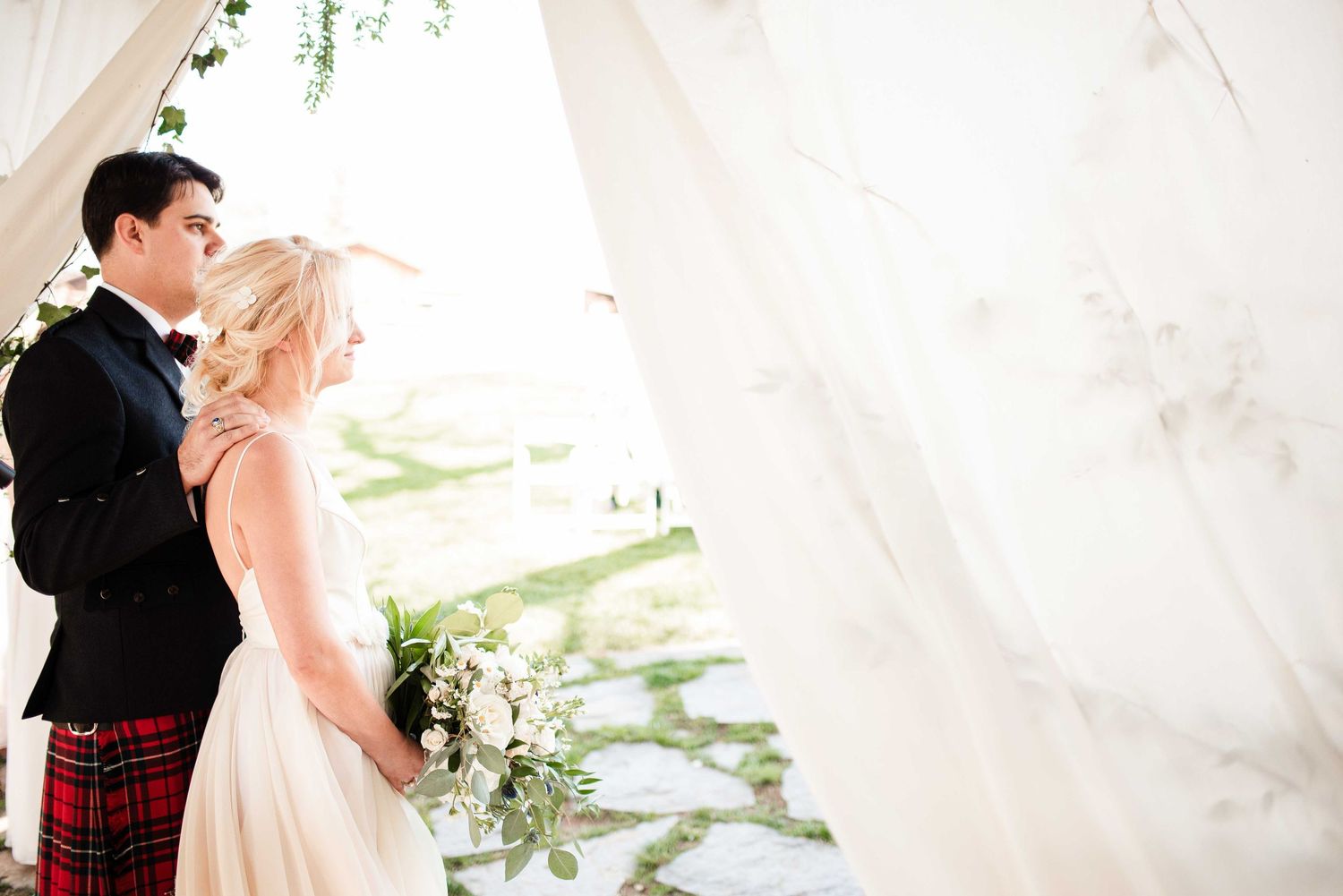Bride and groom stand together under flowing white drapes, groom in tartan kilt, bride holding white bouquet.