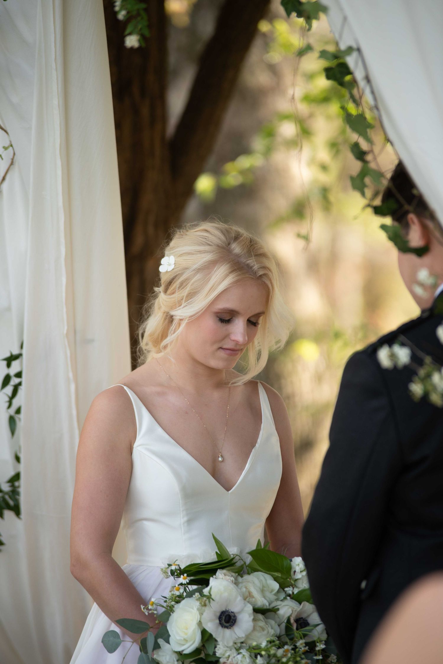 Blonde bride in white gown holds white floral bouquet with anemones at outdoor wedding ceremony.