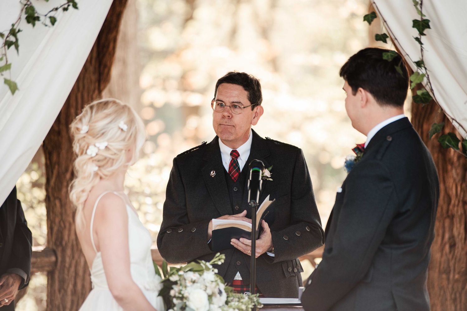 Wedding officiant reads vows as bride and groom stand beneath draped fabric and ivy-covered tree.
