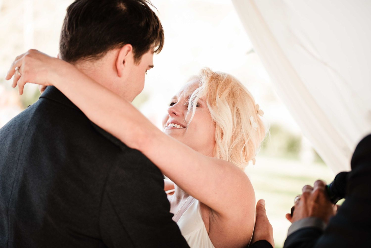 Smiling blonde bride embraces groom in gray suit during romantic first dance at outdoor wedding reception.