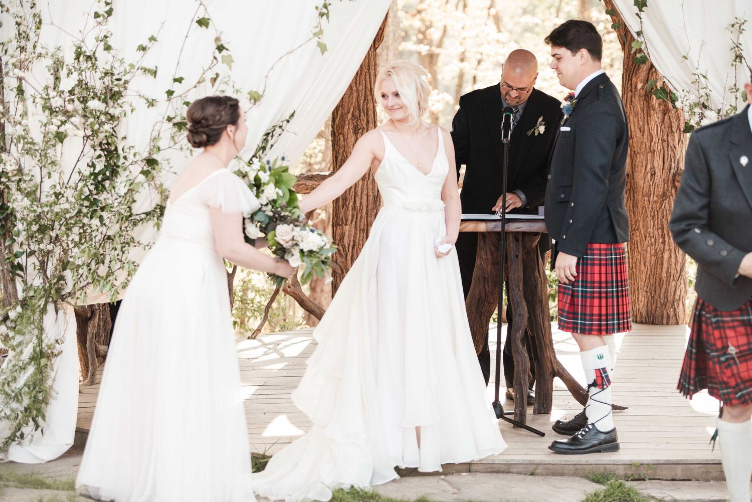 Two brides in white gowns hold hands at outdoor wedding ceremony with kilted groomsman and officiant.