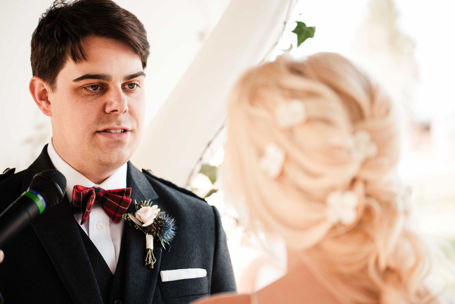 Groom in dark suit with tartan bow tie gazes lovingly at blonde bride during wedding ceremony.