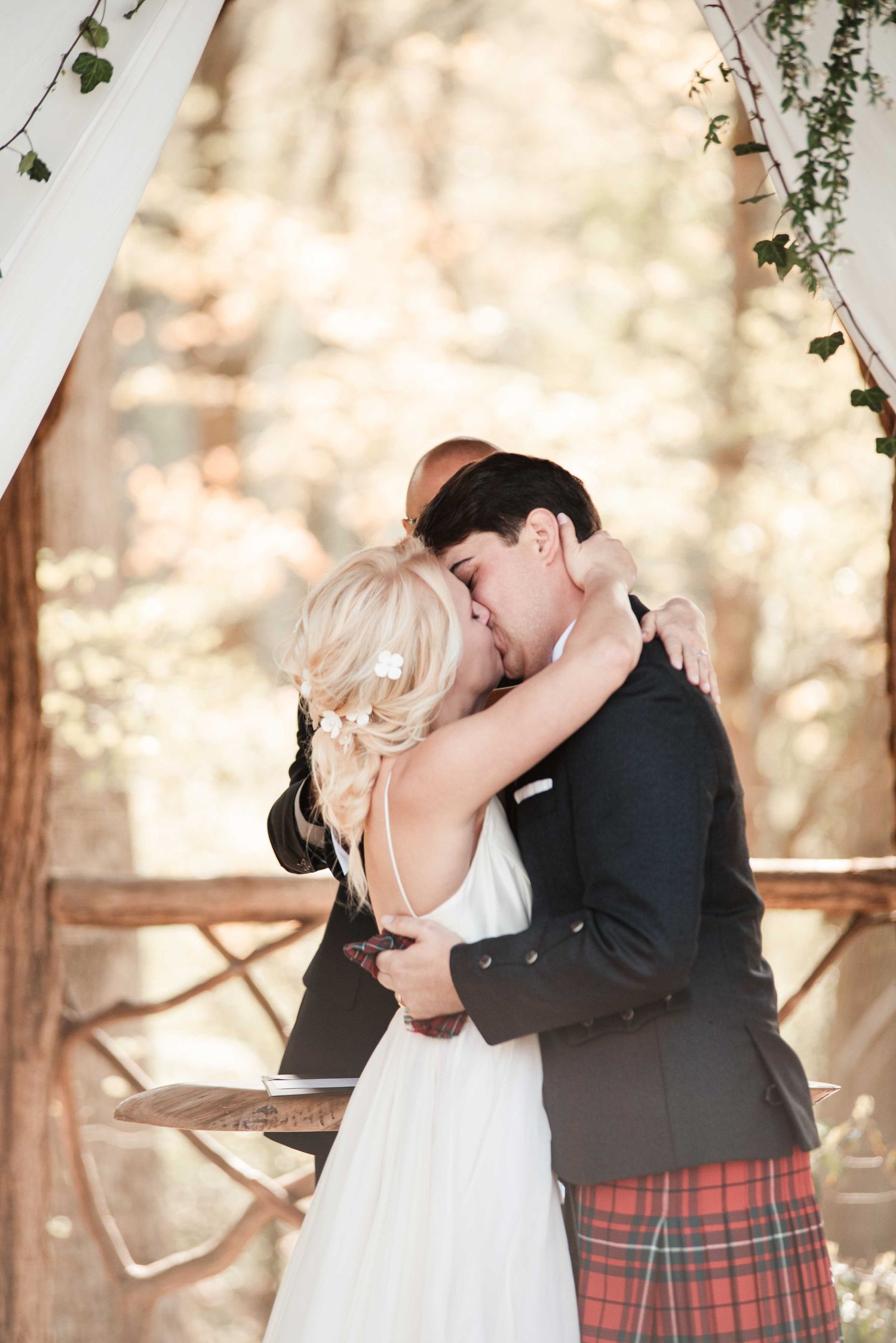 Bride and groom share their first kiss at an outdoor woodland wedding ceremony, groom wearing a red tartan kilt.