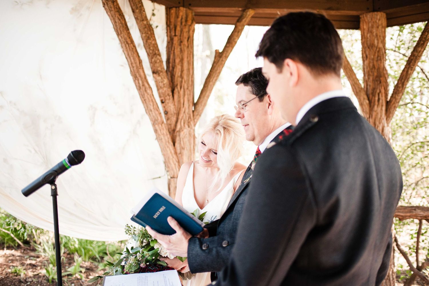 Couple exchanging wedding vows under rustic wooden gazebo with officiant holding blue book at outdoor ceremony.