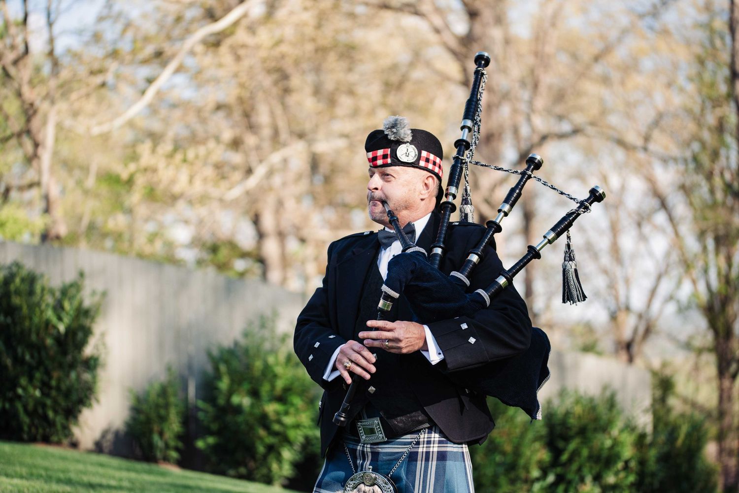 Bagpiper in traditional Scottish attire with kilt and cap performs outdoors in a lush green setting.