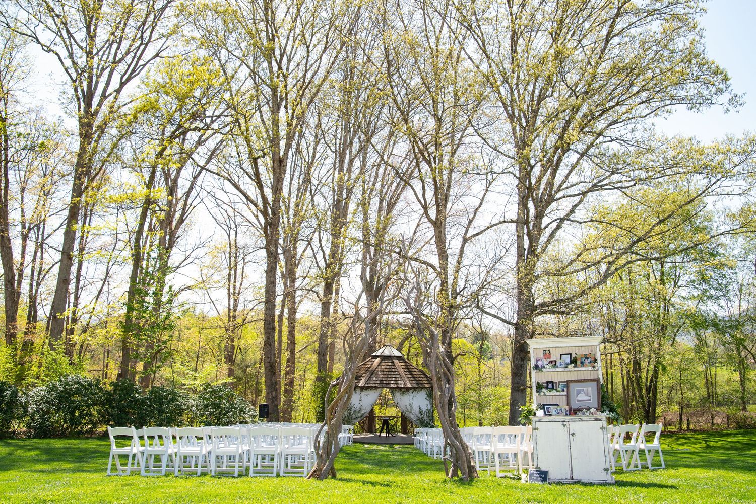 White chairs arranged for outdoor wedding ceremony under tall trees in green meadow.