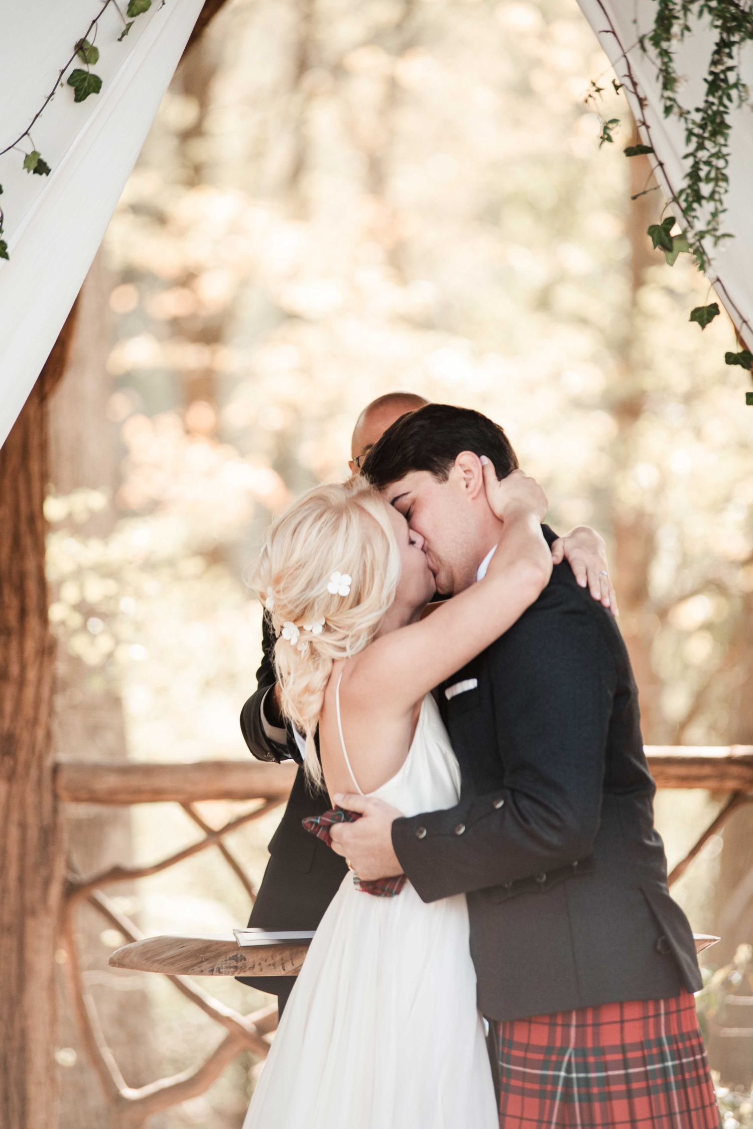Bride and groom sharing first kiss under draped floral arch at outdoor woodland wedding ceremony.
