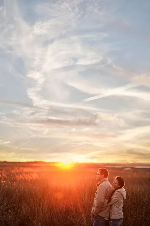 Couple embracing in golden wheat field at sunset with dramatic cloudy sky backdrop.